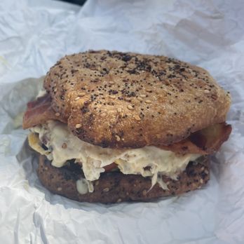 Meta Cafe - The Cait (from the secret menu) - A breakfast sandwich with a multigrain bagel topped with seeds, containing egg, bacon, and cheese. The sandwich is wrapped in white paper, and the background shows a blurred outdoor setting with a vehicle. The image emphasizes the sandwich's ingredients and bread texture. - Sandwiches Near Me - Lambertville, New Jersey