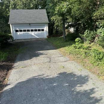Erick Landscaping - A concrete driveway leads to a white garage with a gray shingled roof, situated at the end of the driveway. The garage has a large door with windows near the top. Surrounding the driveway are trees and bushes, providing a green backdrop. Sunlight creates shadows on the driveway, indicating a clear day. - Snow Removal Near Me