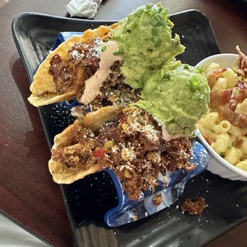 Loading Gastrobrunch - A black rectangular plate holds three tacos filled with seasoned ground beef, topped with cheese and garnishes. Two scoops of guacamole with chopped green onions are placed on top of the tacos. A small bowl of macaroni and cheese with bacon is partially visible on the side. The presentation is on a dark wooden table, with a napkin and utensils nearby. - Breakfast & Brunch Near Me - St Cloud, Florida