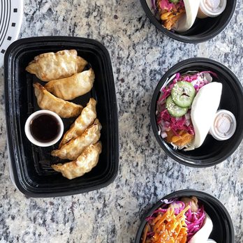 Take a Bao Eats - The image shows three black bowls filled with Asian-inspired salads featuring shredded vegetables, pickles, and sauces, each garnished with sesame seeds and fresh vegetables. A black container holds five fried dumplings served with a small cup of dipping sauce. The items are placed on a speckled granite table, with part of a white circular object visible in the top left corner. - Asian Fusion Near Me - Chicago, Illinois
