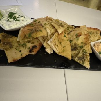 Estiatorio Fili - A black rectangular plate holds sliced flatbread garnished with herbs, accompanied by two small bowls of side dishes—one with cucumber and yogurt, and the other with a creamy dip topped with chopped green onions. - Greek Near Me - Providence, Rhode Island