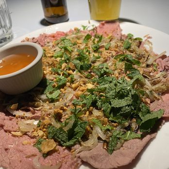 Cafe Da Lat - Sliced Beef Appetizer...simply amazing - Sliced beef is garnished with chopped herbs, onions, and crushed peanuts, accompanied by a small bowl of dipping sauce, with drinks in the background. - Vietnamese Near Me - Albuquerque, New Mexico