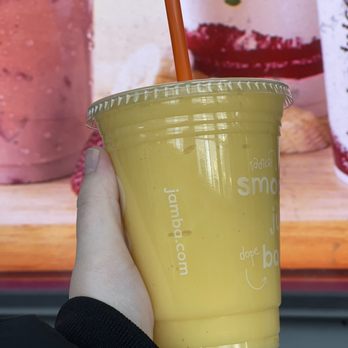 Jamba - A person is holding a yellow smoothie in a clear plastic cup with a matching lid and a brown straw. The background features a colorful, abstract design with pink, red, and green elements. The person's hand is visible, and they are wearing a black sleeve. The cup has white text and graphics, including a website URL and some words, indicating it is from Jamba. - Juice Bars & Smoothies Near Me - Chicago, Illinois