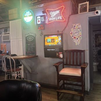 PMO Club - The image shows the interior of a bar or restaurant with wooden flooring and walls decorated with neon signs, including Miller High Life and other beverage advertisements. There is a seating area with chairs, a small table, and a wall-mounted ATM or payment kiosk. A wooden chair with a cushioned seat is positioned near the restroom door, which is open, revealing a toilet inside. The space has a casual, rustic decor with framed pictures and various posters. - Bars Near Me - Lake Villa, Illinois