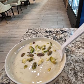 Dera - Rice Pudding - A bowl of rice pudding garnished with chopped pistachios is placed on a marble countertop inside a restaurant with dining tables and chairs. - Pakistani Near Me - Springfield, Virginia