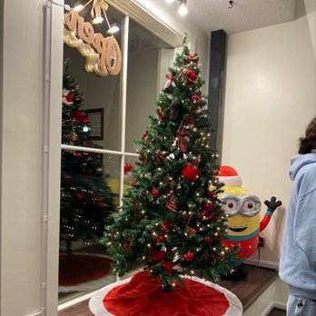 Ming’s Bubble Tea - Inside during the holidays. - An indoor scene features a decorated Christmas tree with red ornaments, ribbons, and string lights. The tree is placed on a red and white tree skirt, positioned near a window that reflects the tree's lights. To the right, a large Minion character inflatable wearing a Santa hat is visible, and part of a person wearing a light blue hoodie is seen on the right side of the image. - Bubble Tea Near Me - Hudson, Ohio
