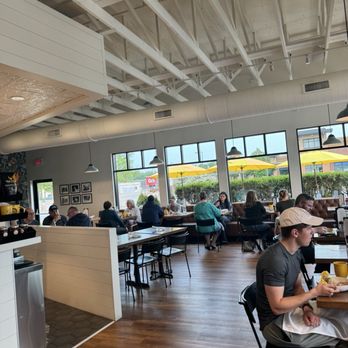 Perk Up Kitchen - The image shows the interior of a restaurant with high ceilings and exposed white beams. Large windows allow natural light to fill the space, revealing outdoor yellow umbrellas. Patrons are seated at various tables, some near the windows, enjoying meals. The decor includes framed pictures on the wall and pendant lighting. The setting appears casual and inviting, with a mix of people dining. - Breakfast & Brunch Near Me - Bloomington, Minnesota