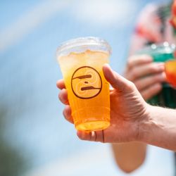 Ziggi's Coffee - Hand holding an iced orange drink in a clear Ziggi's cup outdoors with friends in the background.