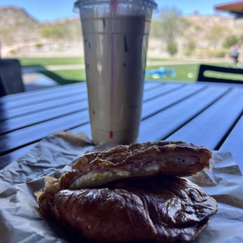 Cafe Bebida - A tall, clear plastic cup filled with iced coffee and a red straw is positioned on a wooden table outdoors. In front of the drink, there is a breakfast sandwich wrapped in paper, featuring a croissant with visible egg, ham, and cheese. The background shows a bright, sunny day with a grassy area, some trees, and distant hills, indicating an outdoor setting. - Coffee & Tea Near Me - Goodyear, Arizona