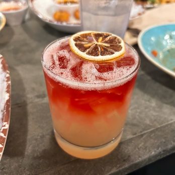 Da Boccery - Tea Bag Cocktail - A cocktail with a gradient from pale yellow at the bottom to red at the top, garnished with a dried lemon slice, served over ice in a clear glass. - Axe Throwing Near Me - Livermore, California