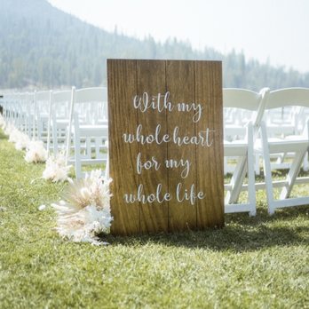 Light and Well - A wooden sign with white cursive lettering reads, 'with my whole heart for my whole life,' and is placed on a well-maintained grassy area. Behind the sign, there are white folding chairs arranged for an outdoor wedding ceremony, with a mountain landscape in the background. The scene is illuminated by natural sunlight, and decorative white floral arrangements are positioned near the sign. - Floral Designers Near Me