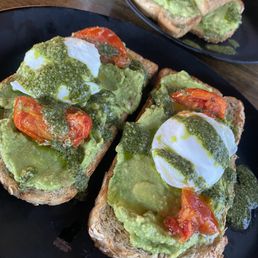 Avocado toast - Two plates of avocado toast are shown, each topped with mashed avocado, poached eggs, cherry tomato slices, and green sauce. The toast is served on black plates, placed on a wooden table. The presentation highlights fresh ingredients and a variety of toppings.