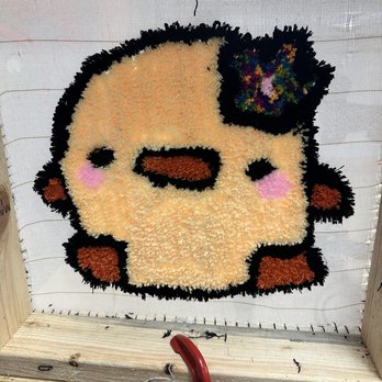 Tuft A Rug - A tufted rug displays a cartoon face with blush cheeks, black eyes, and a nose, with a colorful bow on top, set against a mesh background. - Art Classes Near Me - Aurora, Illinois