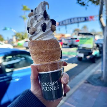Crispy Cones - Cookie à la Mode Crispy Cone - A person holds a soft serve ice cream cone topped with various toppings, including chocolate and possibly cookie pieces, outside on a sunny day. The cone is wrapped in a black sleeve with the logo 'CRISPY CONES.' The background shows parked cars, a blurry sign, trees, and a clear blue sky, indicating an outdoor setting near a shopping or dining area. - Desserts Near Me - Carlsbad, California