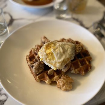 Refinery 51 - Chicken and waffles - A dish of chicken and waffles is served on a white, round plate. The waffles are topped with pieces of fried chicken and a fried egg. The plate is placed on a marble table, with glasses of water and other drinks visible in the background. The setting appears to be a restaurant with a chic ambiance. - Cocktail Bars Near Me - Poughkeepsie, New York