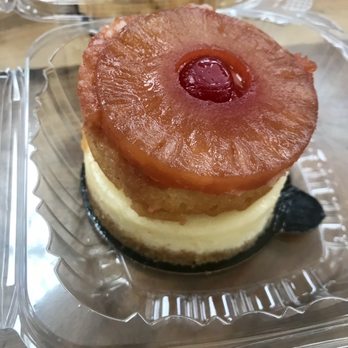 The Cheesecake Cafe - Pineapple upside down cheesecake - A pineapple upside-down cheesecake with a cherry on top, served in a clear plastic container, with a layer of cream cheese filling and a graham cracker crust. - Desserts Near Me - Baltimore, Maryland