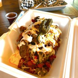 505 potato - A baked potato topped with shredded cheese, a whole jalapeño pepper, and various other toppings such as chopped vegetables and sauces, served in a foam takeout container. In the background, there are checkered paper-wrapped items on a metal tray and a small cup of sauce on a wooden table.