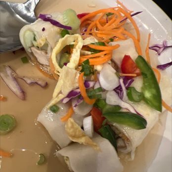 Thai Pavilion - Spring rolls - A plate of fresh spring rolls contains rice paper wrapped around vegetables such as carrots, cucumbers, and lettuce, with herbs and possibly chicken. The rolls are garnished with shredded carrots, sliced cucumbers, and chopped green onions. The dish is accompanied by a creamy, light-colored dipping sauce. The presentation emphasizes the colorful ingredients and traditional Thai or Vietnamese cuisine style. - Thai Near Me - DeKalb, Illinois