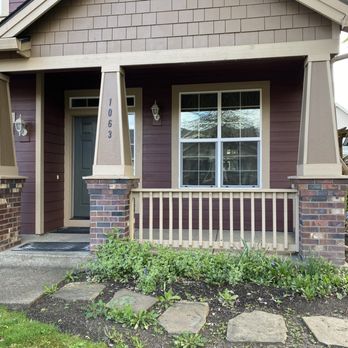 Wild Iris Custom Home Renovations - Before - A house features a porch with brick columns, a gray front door, and a large window. There is a garden with plants and stepping stones in front. - Carpenters Near Me