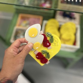Strange Ways - A hand holds a decorative item resembling a fried egg with a white base and yellow yolk, adorned with small cherries and green leaves. The background shows a display of yellow shoes and other colorful items, likely in a gift shop setting. The display case and other merchandise are visible behind the hand, indicating a retail environment focused on unique and indie products. - Accessories Near Me - New Haven, Connecticut
