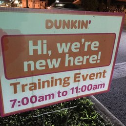 The image shows a signboard placed on the sidewalk near a street, with a background of a road and some buildings. The sign has a white background with pink and brown text, indicating a Dunkin' training event from 7:00 am to 11:00 am. The sign is supported by a metal frame and is positioned among some grass and pavement.