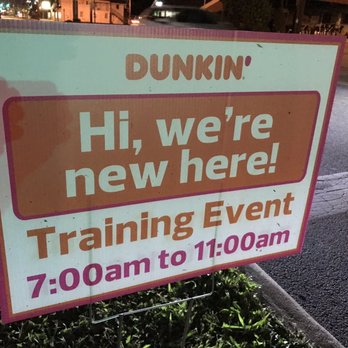 Dunkin' - The image shows a signboard placed on the sidewalk near a street, with a background of a road and some buildings. The sign has a white background with pink and brown text, indicating a Dunkin' training event from 7:00 am to 11:00 am. The sign is supported by a metal frame and is positioned among some grass and pavement. - Breakfast & Brunch Near Me - Columbus, Ohio