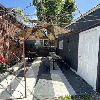 The Vault - Outdoor seating - The image shows an outdoor patio with a concrete floor, enclosed by a brick wall on the left and a painted dark wall on the right. There are two picnic tables with benches, a small round table with chairs, and umbrellas providing shade. Hanging plants are suspended from the umbrellas, and potted flowers are placed near the brick wall. The area is illuminated by sunlight, with a tree visible in the background. - Salad Near Me - Gilbert, Iowa