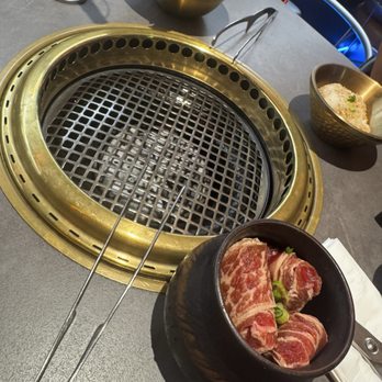 Y Space BBQ - The image shows a table at a Korean barbecue restaurant featuring a built-in grill in the center. There is a bowl of raw meat, likely beef, with green onions, a bowl of rice, and a bowl of fresh lettuce. The table surface is gray, and the grill is metallic with a gold-colored rim. The setting suggests a dining experience where customers cook their own meat and vegetables at the table. - Barbeque Near Me - Flushing, New York