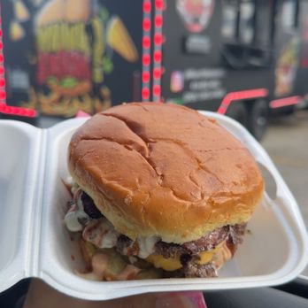Munch Mobile - Munch Monster Smashburger - A hand holds a foam container with a smashed burger topped with cheese, bacon, and sauce. The background features a black food truck decorated with colorful graphics, including a burger and a crane, with red lights outlining its edges. The scene suggests street-side dining at a food truck event. - Burgers Near Me - Milwaukee, Wisconsin