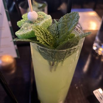Trevor's - WMPO special drink - A tall, clear glass holds a light green cocktail, garnished with fresh mint leaves, lime slices, and a decorative pick with a small golf ball on top. The drink appears to be served in a dimly lit setting, possibly a bar or restaurant. - Pizza Near Me - Phoenix, Arizona