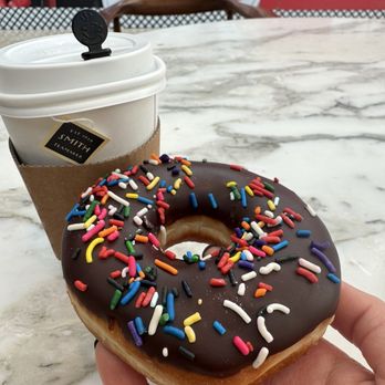 Glaze Donuts - A hand holds a donut topped with chocolate glaze and multicolored sprinkles, positioned on a marble table. In the background, there is a coffee cup with a sleeve and lid, and a blurred view of sushi plates on a wall. The setting appears to be a cafe or restaurant with a casual dining environment. - Donuts Near Me - Paramus, New Jersey