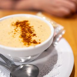 Cream of Crab Soup - A close-up of a white ceramic bowl filled with cream of crab soup, garnished with a line of spices or paprika. The bowl is placed on a white plate with a paper doily, accompanied by two silver spoons. The background shows a wooden table and a blurred hand, indicating a dining setting at Koco's Pub in Baltimore, MD.
