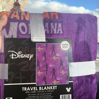 Five Below - Blanket - A purple Disney Hannah Montana-themed travel blanket, labeled as super soft, is being held up inside a vehicle. The blanket features silhouettes of Hannah Montana in various poses, along with Hannah Montana branding. The background shows a bright blue sky with some clouds visible through the windshield, and a rearview mirror with a hanging air freshener is also visible. - Discount Store Near Me - Albuquerque, New Mexico