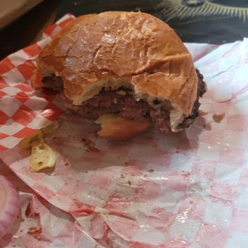 101 Drive-In - The burger we didn't ask for - A partially eaten burger with a soft bun and beef patty rests on red and white checkered paper. The burger appears to have some condiments and toppings, with crumbs and remnants of the bun and meat visible. The background includes a dark surface and a glass, indicating a casual dining setting. - Burgers Near Me - Willits, California