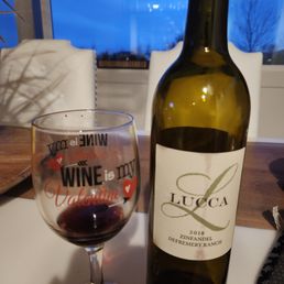 Lucca wine for V day in Idaho!  We love their wine!  Our only wine club we have shipped to Idaho! - A wine bottle labeled 'Lucca 2018 Zinfandel Defremery Ranch' is positioned next to a glass containing red wine. The setting appears to be indoors, with a window revealing a blue evening sky and some trees outside. The table surface is wooden, and there are chairs and a plant partially visible in the background.