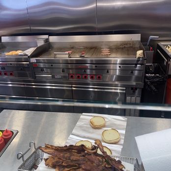 Five Guys - Fresh burgers - The image shows a commercial kitchen with a stainless steel griddle in the background, where some pieces of meat are cooking. In the foreground, there are burger buns on a paper towel and a basket filled with cooked bacon strips on a stainless steel counter. The kitchen appears clean and organized, with cooking tools and supplies visible. - Burgers Near Me - Catonsville, Maryland