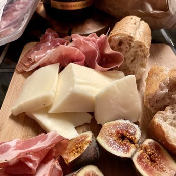 The Cheeseboard Collective - Teleme cheese - A cheese and charcuterie board featuring various cheeses, prosciutto, fresh figs, bread, and a bottle of Susumanello wine. - Bakeries Near Me - Berkeley, California