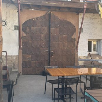 305 Fire - Outdoor patio area with metal tables and chairs, featuring a large rusted metal door with a decorative shape, against a stone and stucco wall. - Pizza Near Me - Gallup, New Mexico