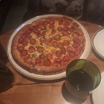 Basso - Pepperoni pizza - A pepperoni pizza with a golden crust, topped with slices of pepperoni, melted cheese, and sprinkled green herbs, is placed on a white plate on a wooden table. Surrounding the pizza are glasses of dark soda, and part of a person's arm and torso are visible in the background, indicating a dining setting. - Bars Near Me - Saint Louis, Missouri