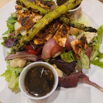 Pasta E Basta - Roasted Garlic Chicken Salad ! - The image shows a grilled chicken salad on a white plate, including mixed greens, roasted asparagus, and grilled chicken pieces with grill marks. Two small bowls of dark dressing or sauce are placed on the plate, one near the top and one near the bottom. The salad is presented on a wooden table. - Italian Near Me - Akron, Ohio