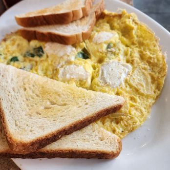 The Pantry - Frittata - A white plate contains scrambled eggs with visible green herbs and white cheese chunks. Three slices of toasted bread are placed on top of the eggs. The plate is on a brown countertop, with a metal cup and a condiment bottle visible in the background. - Diners Near Me - Westerly, Rhode Island