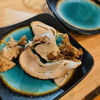 Speakeasy Miso - Boiled bacon. Not pork belly - Slices of boiled bacon are placed on a black and blue ceramic plate, with some pieces appearing tender and fatty. An empty matching plate is positioned behind the main plate. The setting is on a wooden table, suggesting a restaurant environment. The plates have a distinctive blue center with a black border, and the background includes a partially visible paper bag and a glass. - Ramen Near Me - TROY, Ohio