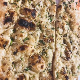 Garlic Naan - The image shows two pieces of garlic naan bread with herbs, featuring a golden-brown, slightly charred surface. The bread appears soft with visible garlic and chopped herbs sprinkled on top. The naan is placed on a surface with some crumpled foil visible in the background, indicating it is freshly baked and ready to serve.