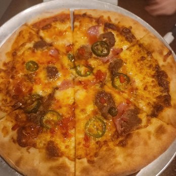 Fire Slice Pizzeria - Honey Bomb Pizza - A pizza topped with jalapeños, sausage, and ham, served on a round metal tray, with a small plate of leftover food in the background. - Pizza Near Me - Amarillo, Texas