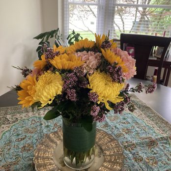 Send Smiles - Bouquet of Flowers - A bouquet of yellow, pink, and purple flowers in a glass vase sits on a decorative tablecloth, with a window and chairs in the background. - Florists Near Me - Chantilly, Virginia