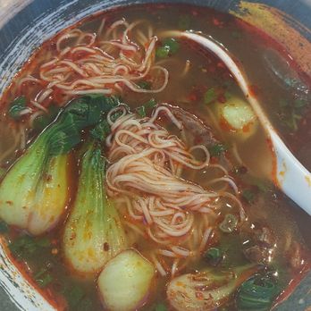 Juicy Dumplings - Beef soup with noodles - A ceramic bowl contains noodle soup with broth, green onions, bok choy, and noodles. A white soup spoon rests inside the bowl. The table surface is white, and there are chopsticks, a small dish, and a spoon visible in the background. - Chinese Near Me - Euless, Texas