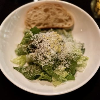 Bocca - Bocca Caesar - A white bowl contains a Caesar salad with chopped romaine lettuce, topped with grated cheese. A slice of bread is placed at the back of the bowl. The setting appears to be in a restaurant environment, with a dark background and some blurred objects. The salad is presented simply, highlighting fresh greens and cheese. - Italian Near Me - Indianapolis, Indiana