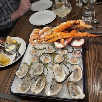 Roost - Seafood platter - A seafood platter displays oysters on the half shell, arranged on ice with lemon wedges and dipping sauces. The platter also includes cooked shrimp, crab legs, and lobster claws, presented in a restaurant setting with glasses of water and wine on the table. The presentation emphasizes fresh shellfish and seafood variety. - New American Near Me - Sparkill, New York
