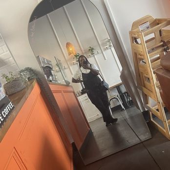 Doughfee - A woman is taking a mirror selfie in a coffee shop with orange and white interior design, shelves with plants, and a cozy atmosphere. - Coffee & Tea Near Me - Jacksonville, Florida