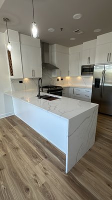Condo Kitchen Remodel - For this kitchen remodel, we transformed the space by painting the existing cabinets for a fresh, updated look. We replaced the old sink and faucet with modern fixtures, and upgraded the countertops from granite to stunning quartz featuring a waterfall edge. The same quartz was also installed seamlessly across the backsplash, creating a clean, elegant, and cohesive design.